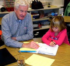 : Bob Buchanan, a retired pediatrician and executive, helps an Angell Elementary student with her math workbook. Buchanan often uses a glass full of coins – from the United States as well as around the world – to bring home math lessons for his pupils at Angell Elementary.  