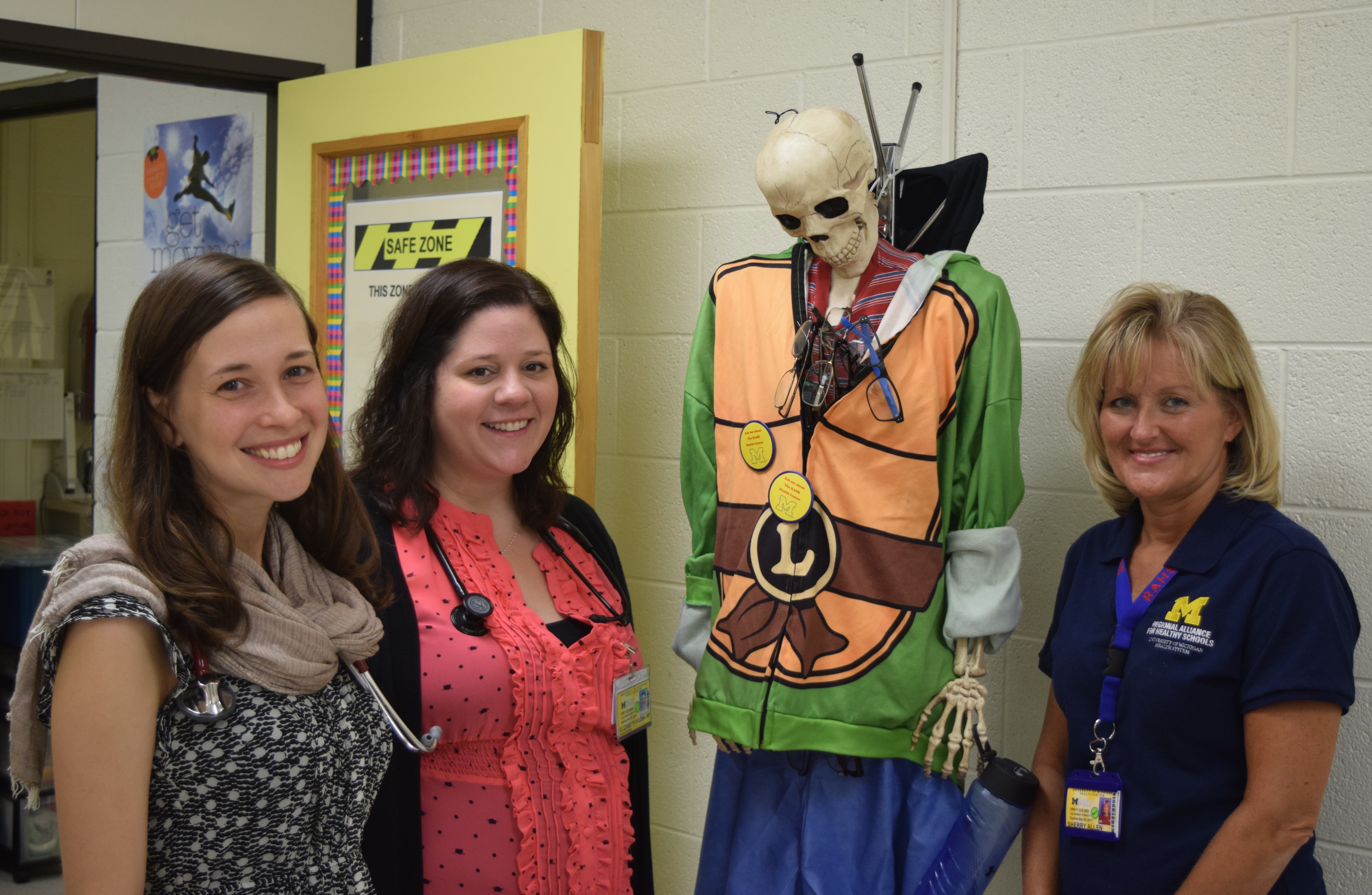 3 staff members at the University of Michigan RAHS Clinic at Scarlett Middle School stand next to a skeleton wearing a Halloween costume