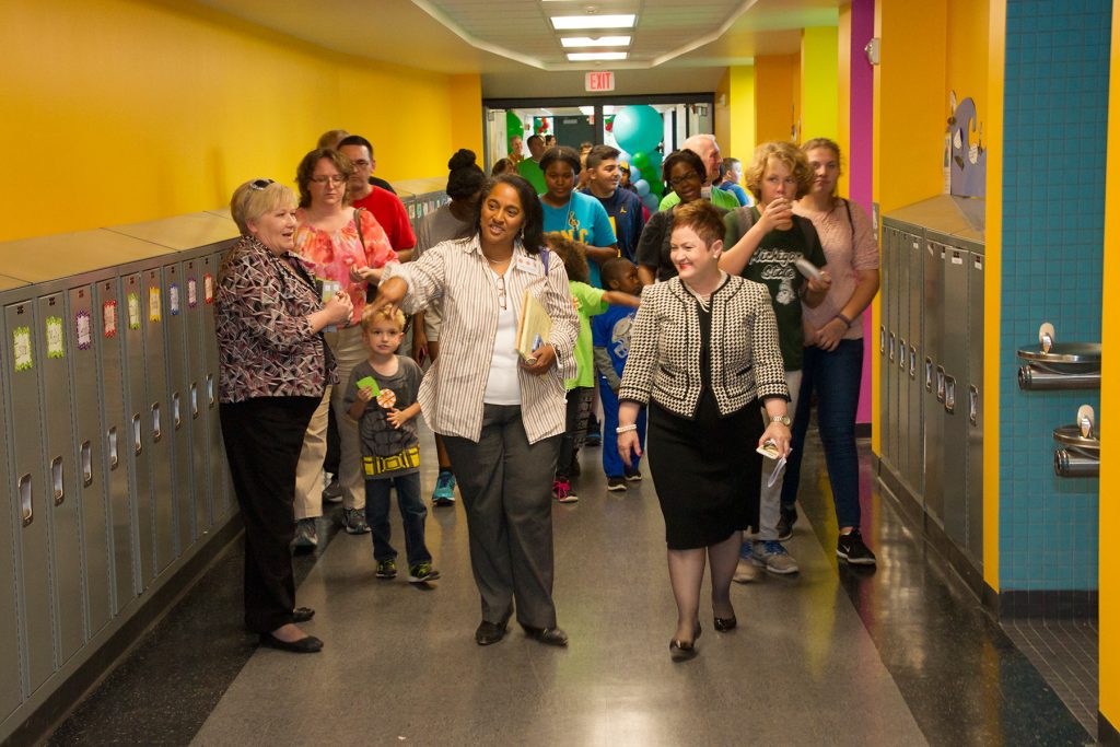 A crowd of people walk down a brightly painted elementary school hallway.