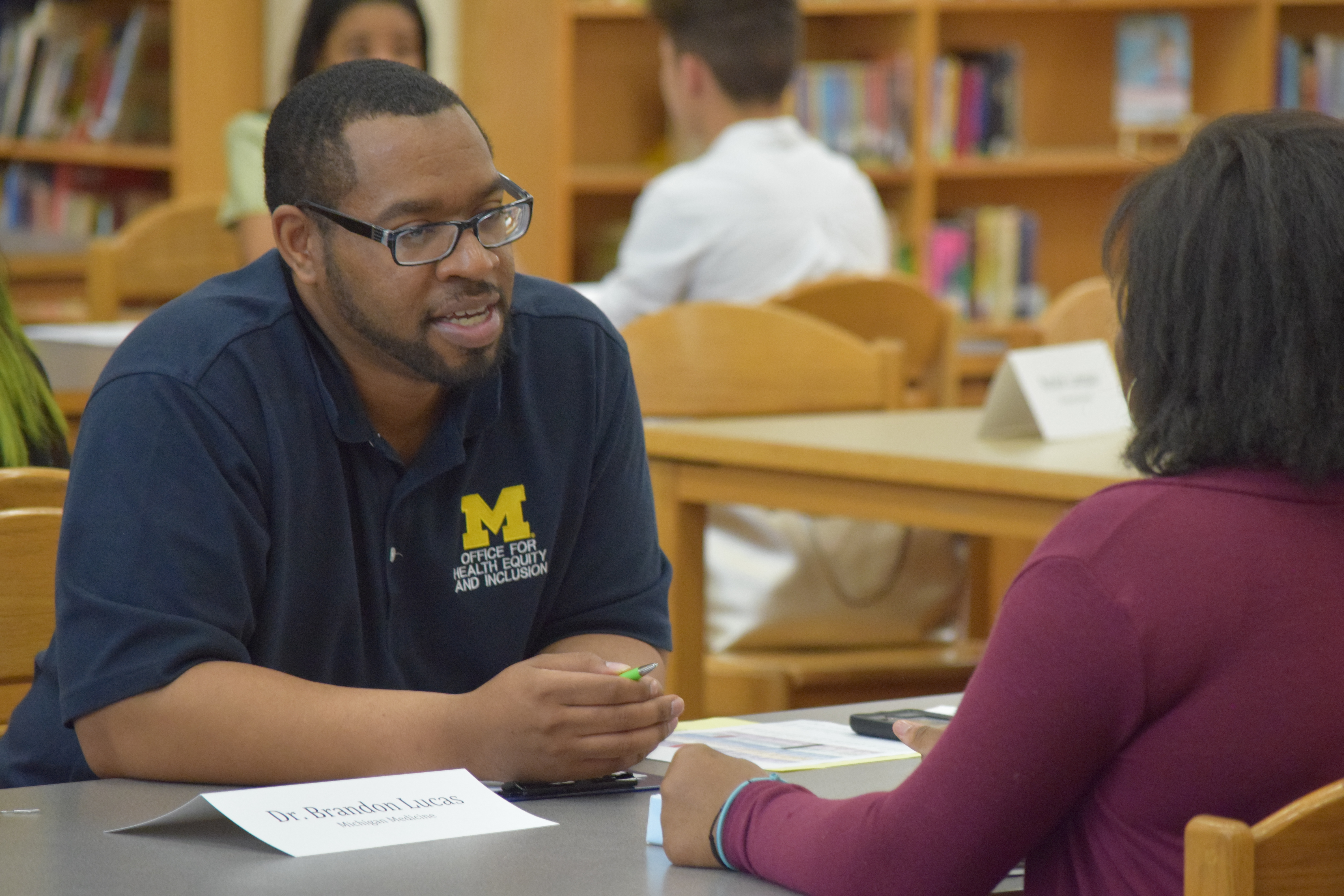 An African-American male wearing a blue shirt sits at a table interviewing an 8th grade student