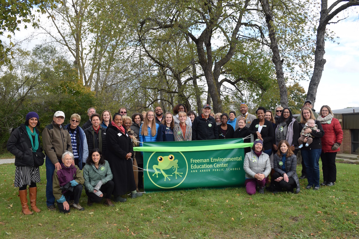 A group of about 50 students, community members, members of the Board of Education, and AAPS teachers and leaders stand behind a ribbon and the Freeman Environmental Education Center's banner.