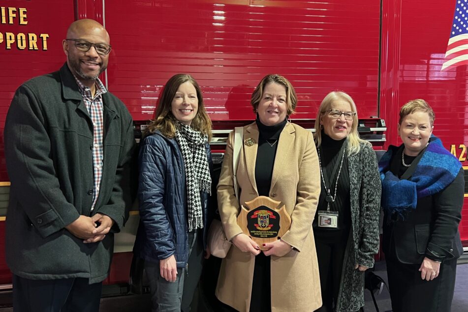 John Randle, Jenna Bacolor, Liz Margolis, Hanna Melangton, and Jeanice Swift stand in front of a fire truck as Margolis holds a plaque honoring her for emergency preparedness work.