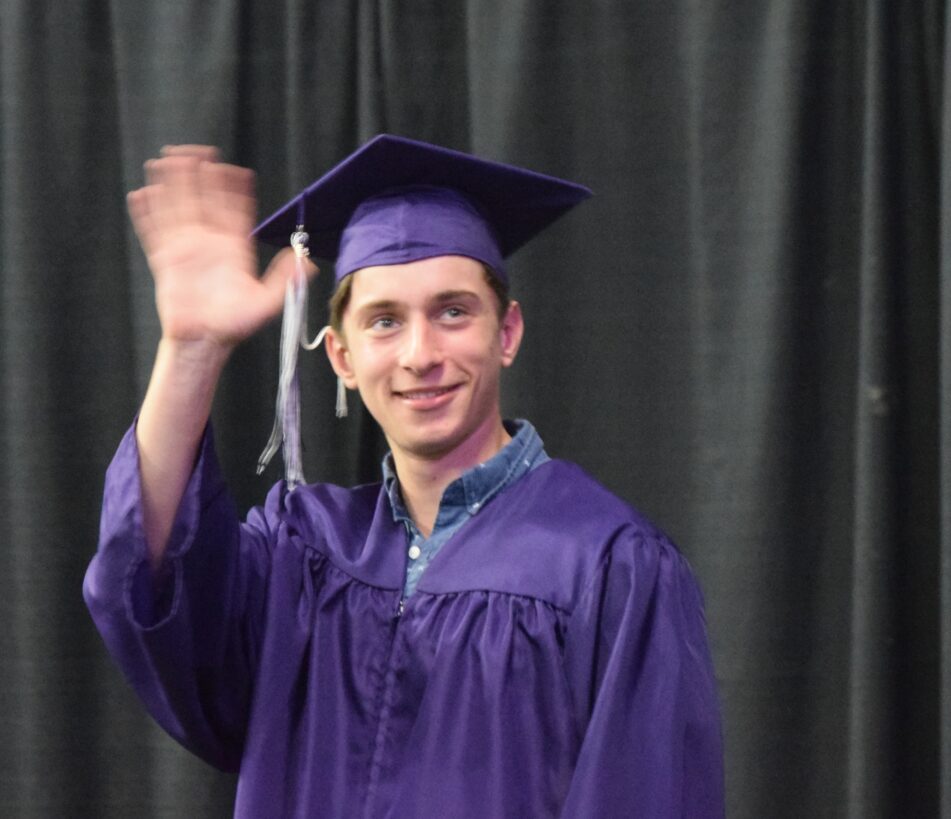 A male Pioneer student in purple cap and gown waves.