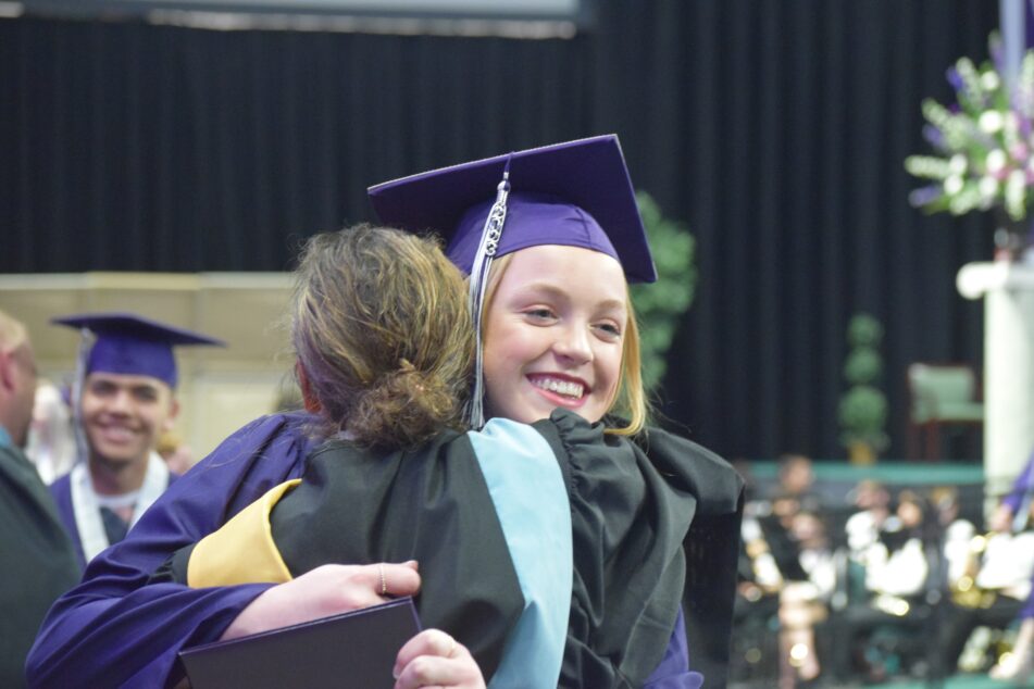 A female Pioneer student in cap and gown hugs a teacher after receiving her diploma.