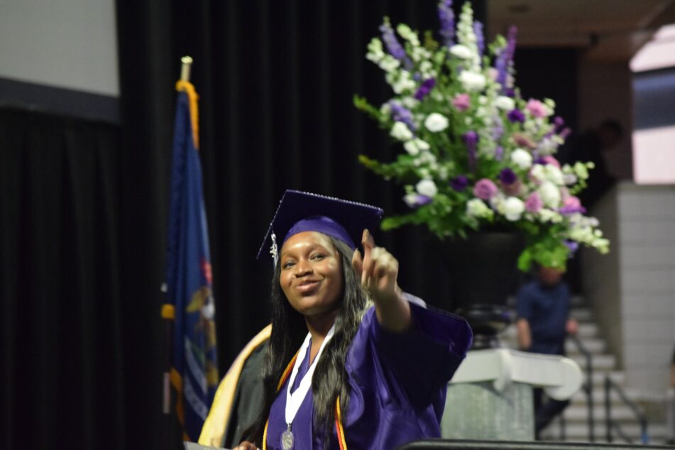 A female Pioneer student in cap and gown points as she crosses the stage after receiving her diploma.