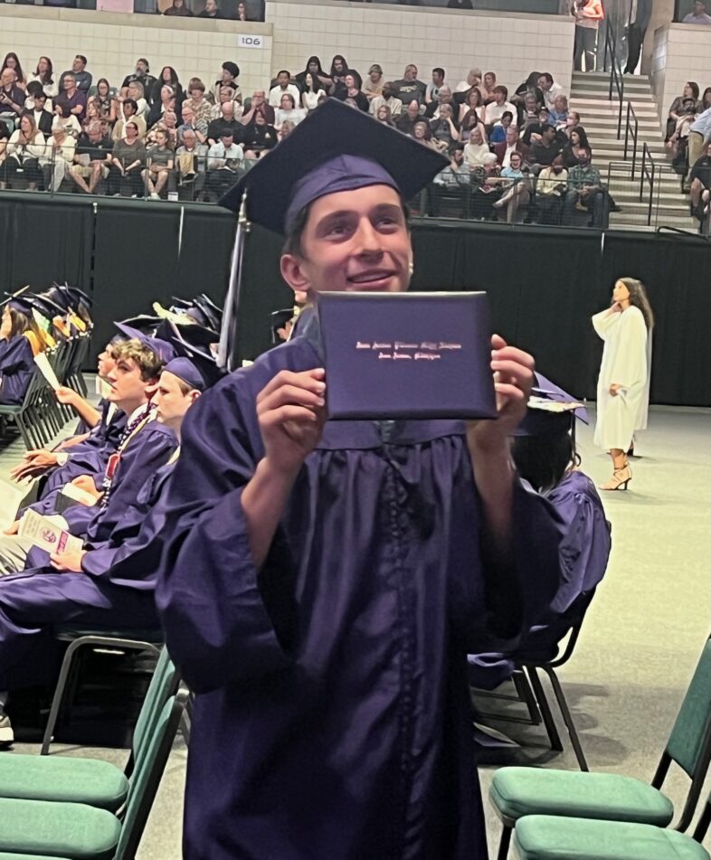 A male Pioneer student in purple cap and gown holds up his diploma to show family in the stands.