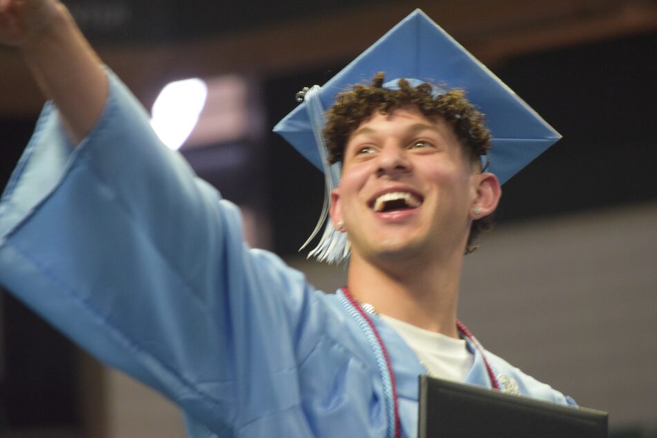 A male Skyline grad in his cap and gown with a big smile points into the crowd after receiving his diploma.