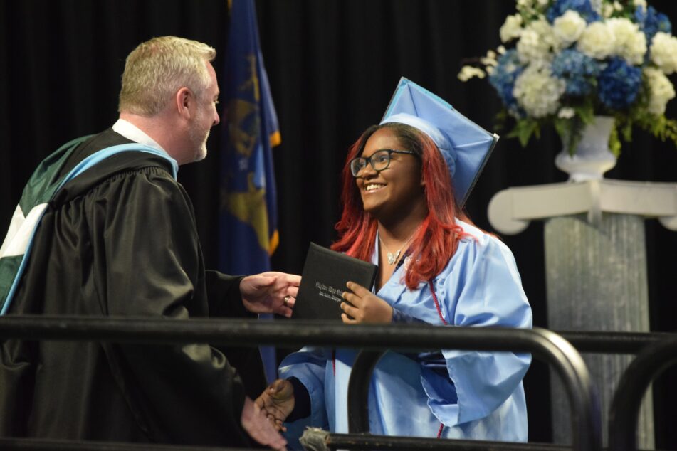 A female Skyline student in cap and gown receives her diploma from Principal Cory McElmeel.
