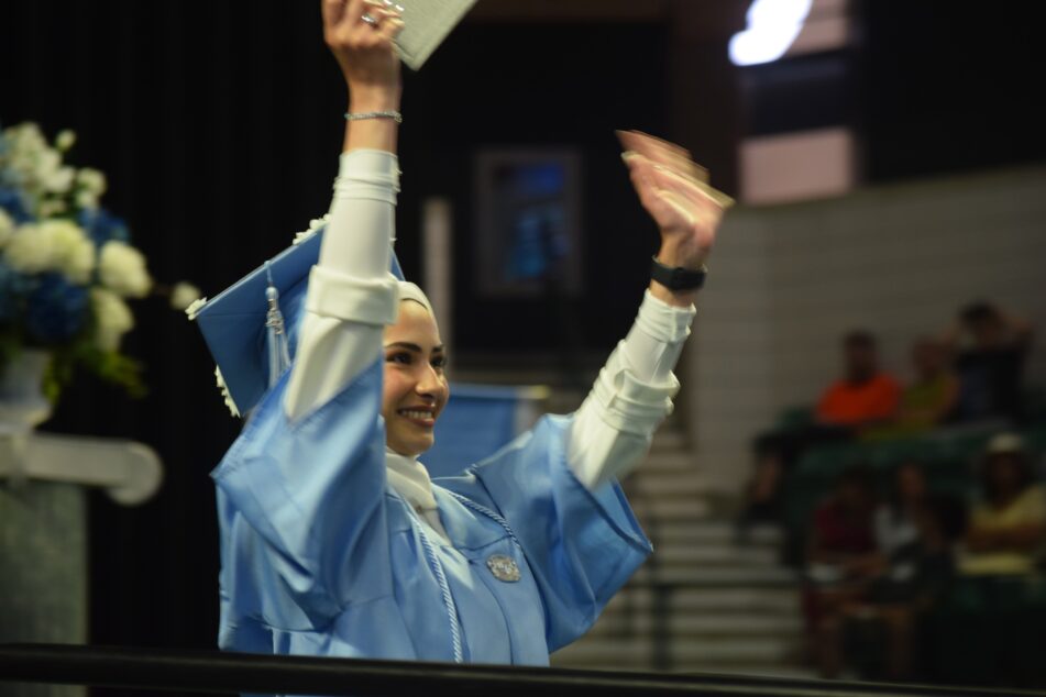 Woman in Skyline cap and gown and white hijab holds up her diploma in one hand and waves with the other.