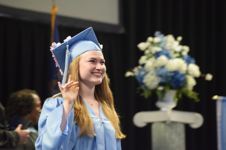 A female Skyline student in blue cap and gown waves after receiving her diploma.