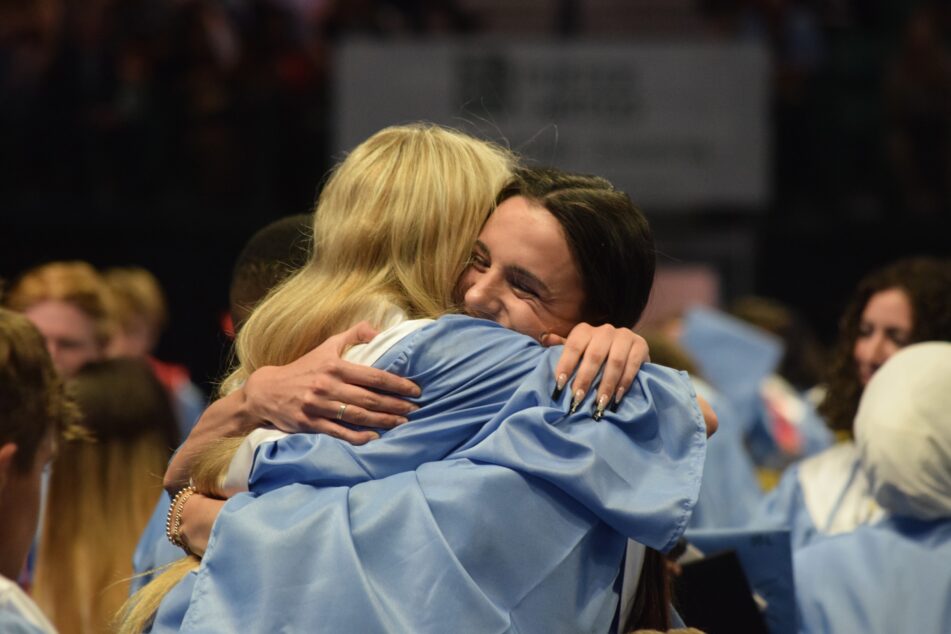 A pair of Skyline students in gowns hug after receiving diplomas and throwing their caps in celebration.