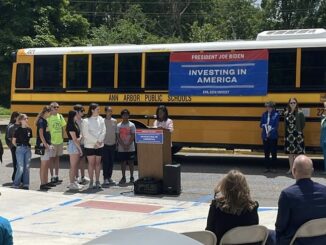 Interim Superintendent Jazz Parks speaks at a lectern in front of an AAPS electric bus, with Clague students on one side of her, and White House Advisors, EPA leaders and elected officials on the other side.