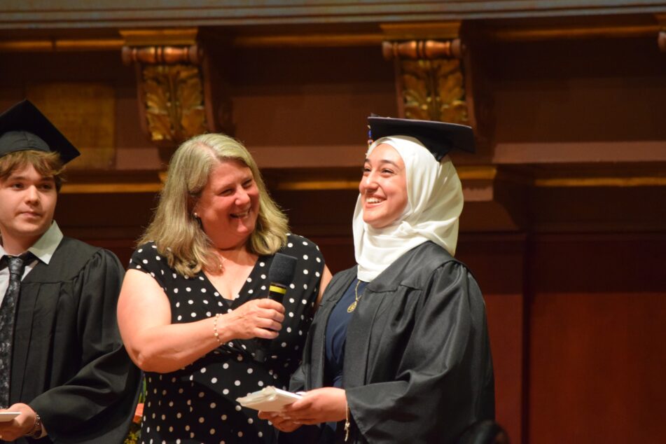 A female in cap and gown and a hijab laughs as she starts her remarks while her former leader and another grad look on.