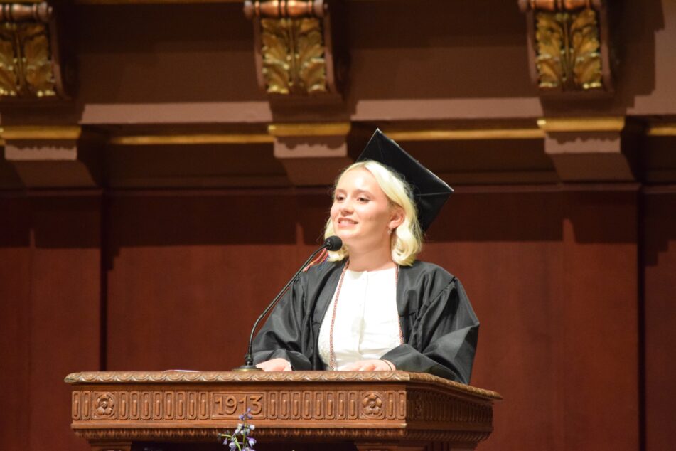Claire Lewis in a open graudation gown with a cap reads her poem at a lectern.