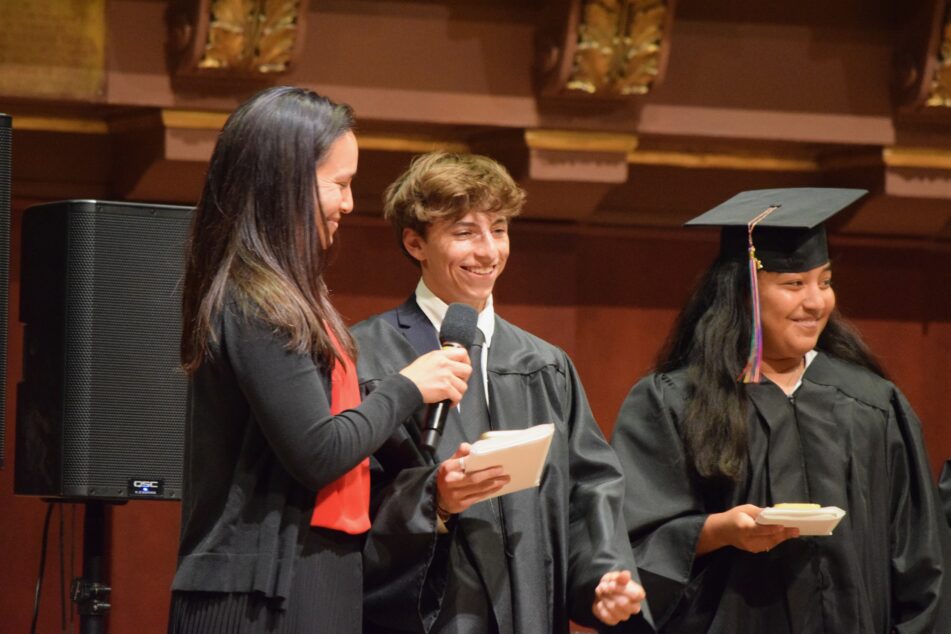 A Community graduate in a graduation gown shares his remark while his former leader and a female in cap and gown listen.