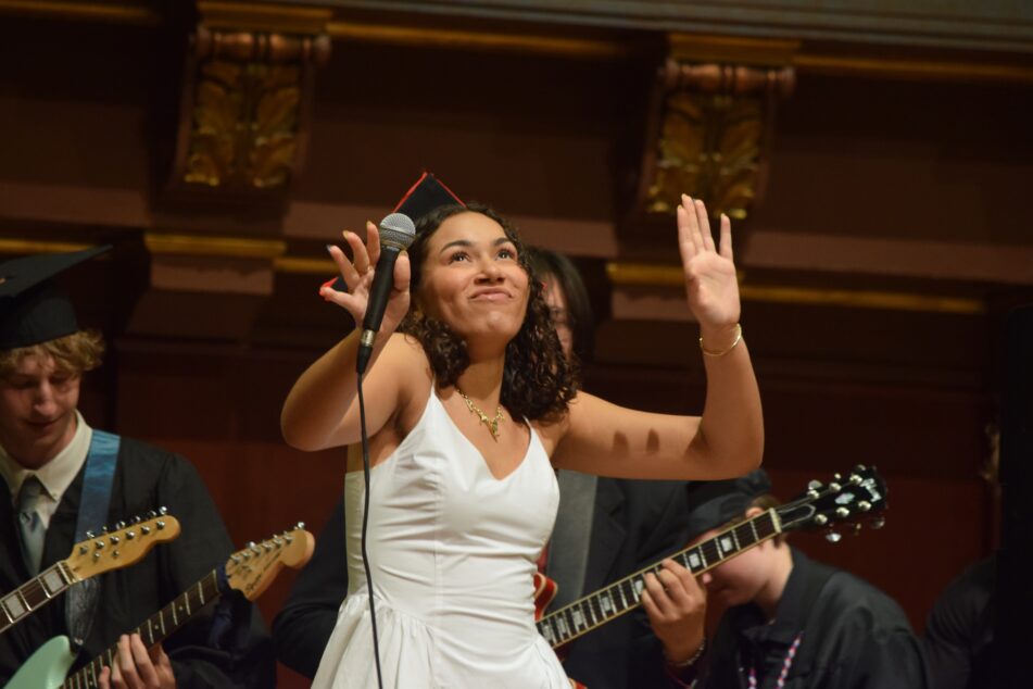 Woman in a white dress and a cap sings on stage with band in the background.
