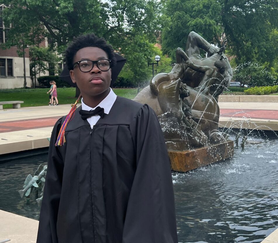A male Community grad in cap and gown looks towards the future in front of a fountain outside before the ceremony.