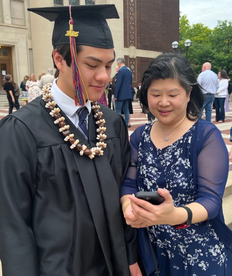 A Community grad in cap and gown and his mother look at a photo on her phone.
