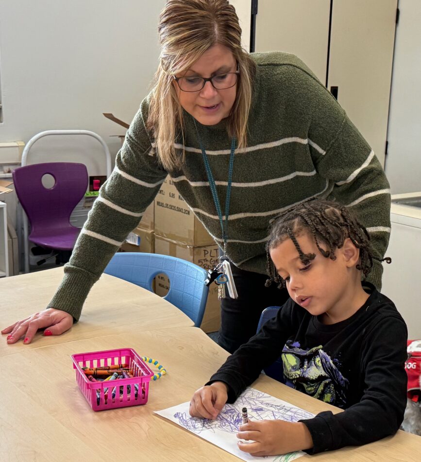 An Angell teacher stands helping a young student who is drawing on a piece of paper.