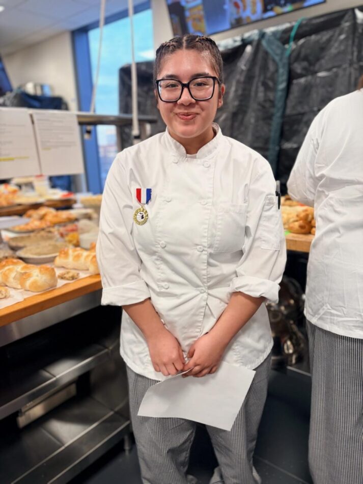 Huron's Liahni Quijada in a chef's smock in a kitchen with a medal pinned to her for winning the baking competition.