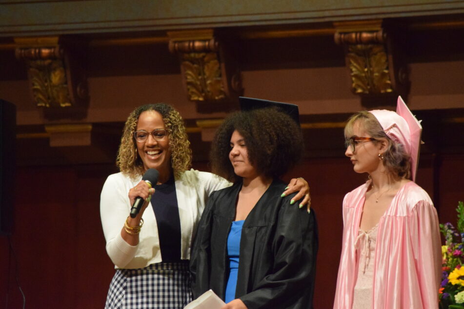A Community grad gets ready to give her speech while another one is next to her and the forum leader is on the other side holding the microphone.